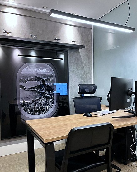 Parede do escritório com um painel em forma de janela de avião mostrando a vista do Rio de Janeiro, e miniaturas de aeronaves na prateleira acima.