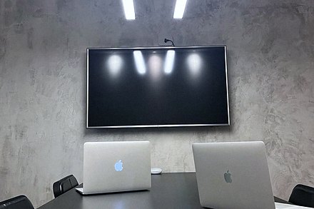 Mesa de reunião vista de frente, com dois notebooks abertos e o televisor de apoio ao fundo.