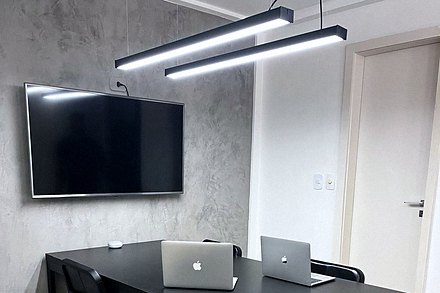 Sala de reunião do escritório, com mesa preta, cadeiras, televisor na parede de concreto e luminárias lineares.